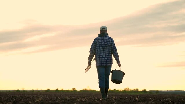 Agricultural industry. Farmer in boots walks across field. Farmer working in field, sunset. Agricultural business concept. Owner of farm with shovel, bucket in hand, go along plowed field. Plantation