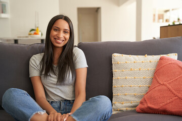 Obraz premium Happy young biracial woman in casual clothing smiling while sitting on sofa in living room at home