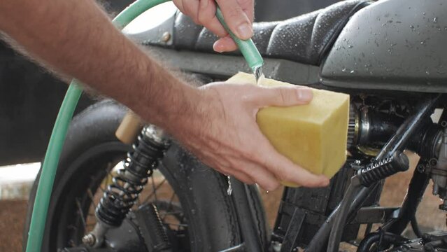 A man washes a custom motorcycle with a sponge and water from a hose. The steady shot shows the cafe racer and the rider squeezing the sponge and water running down. The bike is the background.