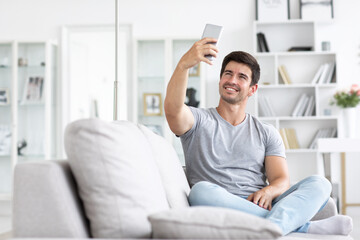 Young handsome happy man talking on a video call or takes a selfie on sofa in living room.