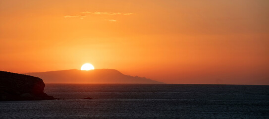 Sunset over Greek island Cyclades Greece. Golden sun hides behind hill silhouette. Calm sea. Banner