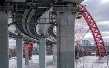 Naklejka premium View of concrete supports of overpass in frozen river and red cable stayed bridge at winter.