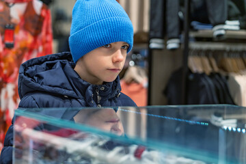 Kid boy stands in shopping mall among clothes and looks at showcase. Teenager chooses new outerwear in clothing store. Shopping and sales concept.