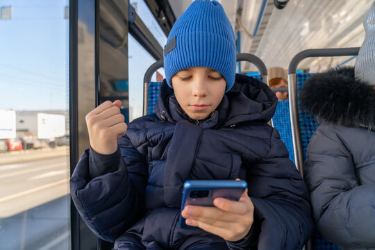 Boy Sits At Window And Looks With Interest At Screen Of Smartphone. Happy Caucasian Kid Using Cell Playing Mobile Games Online On Smartphone Connected To Public Wifi Sitting On Seat In City Tram.