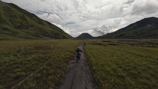 Aerial view biker riding on moto enduro path between volcano mountain landscape extreme adventure. FPV drone follow shot motorbike motocross speed racing power race trip at rock scenery cloudy sky 4k