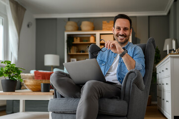 Portrait of confident cheerful male freelancer holding laptop and sitting with laptop on armchair in home office Smiling young businessman checking e-mails over wireless computer while working at home