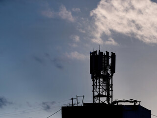Silhouette of telecommunication tower with microwave panel antennas, remote transmitter units, wireless technology, GSM, 5G, optic fibers on roof of building in blue sky with clouds.