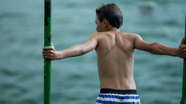 Back of wet young boy standing by pier lake. Contemplative preteen kid boy stands in outdoor nature at beach summer vacations
