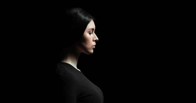 Woman stands in profile against a black background. She slowly and dramatically lifts her head upwards and gazes at the light with a serious and thoughtful expression.