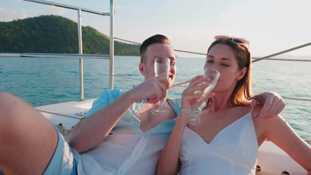 Caucasian romantic couple drinking champagne on deck of yacht while enjoy honeymoon trip. Attractive man and woman party, celebrating anniversary journey trip embrace while holding cocktails on sunset