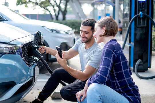 White man and girl fill electrical car green energy , while looking at smartphone with smile of happiness while waiting for refluel.