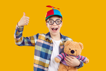 Fototapeta Happy funny boy wearing thick glasses and cap with propeller. Portrait of joyful teenage boy in plaid shirt holding teddy bear toy standing on yellow studio background showing thumb up obraz