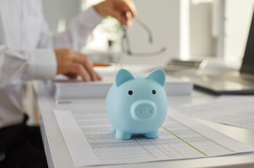 Blue piggy bank on the table with a businessman accountant in background working at home or at office with documents, tables and charts making calculations. Investment and savings concept.
