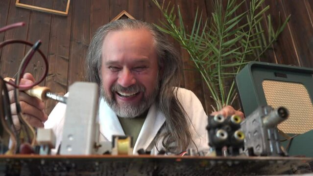 Portrait of an elderly long-haired man, crazy craftsman holds hot soldering iron in his hands and laughs at camera