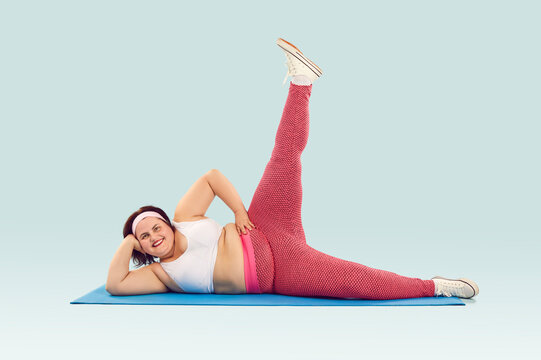 Happy, fat woman enjoying fitness exercise workout. Cheerful, funny, smiling, overweight, big, large woman in sportswear lying on sports mat and raising one leg up isolated on light blue background