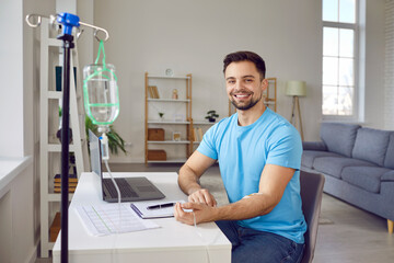 Portrait of happy, smiling, young man patient receiving intravenous vitamin infusion through sterile IV line in his arm while sitting at desk with laptop computer at home. Vitamin therapy concept