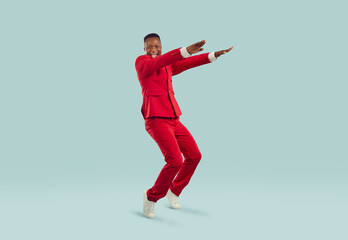 Fototapeta na wymiar Cheerful excited stylish african american man having fun on pastel gray background. Happy fashionable man in red suit dances funny standing on his toes with outstretched arms. Full length.