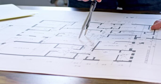Engineer draws circle using dividers on latest floor plan of building project. Workman draws standing near table with drawing and calculator
