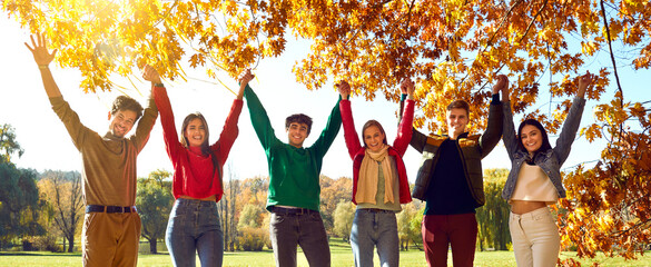 Group joyful friends enjoying good weather, breathing fresh air, having fun in autumn park. Six happy young people holding up hands standing in row in bright sunshine under yellow maple tree. Sunflare