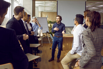 Serious young man business trainer talking to group of people in office standing around him and listening. Office staff, entrepreneurs on corporative training, team building seminar or master class. - obrazy, fototapety, plakaty