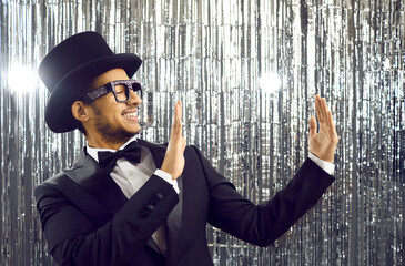 Smiling young latin american in black tuxedo holding in the hands of empty space on shiny silver background. Positive elegant guy in glasses, classic suit, bow tie and top hat looks at the copy space.