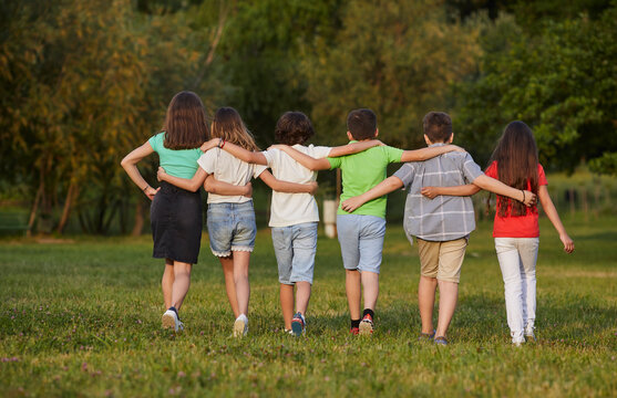 View From Back Boys And Girls Walk Together On Grass Hugging Each Other Walking In Casual Summer Clothes With Children From Elementary School Located On Green Lawn Next To Trees. Schoolchildren, Kids