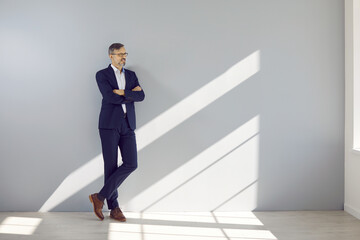 Middle aged handsome businessman leaning against gray wall. Full length portrait of attractive mature man in eyeglasses and formal wear standing with folded hands and looking at window