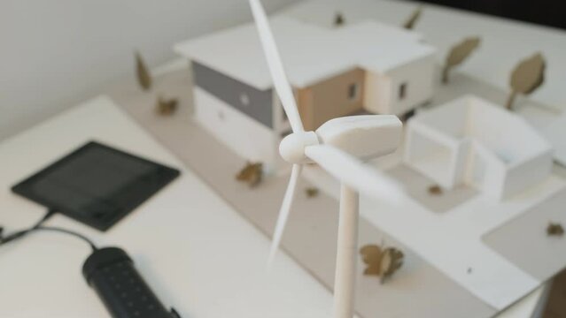Close-up shot of white small scale architectural model of environmentally sustainable home, on table in office of renewable energy research company, and toy wind turbine with rotating blades