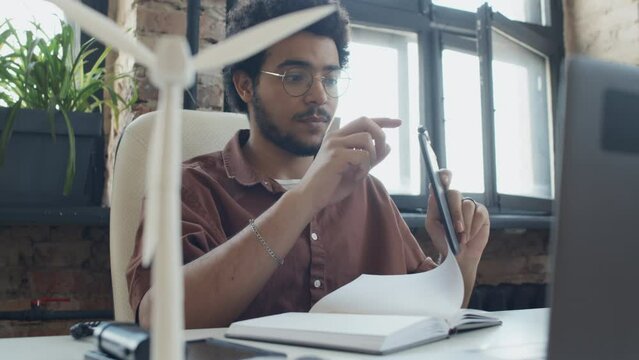 Medium arc shot of young Arab male product designer sitting at desk, drawing in notebook, then picking up graphic tablet, and wind turbine model with portable solar battery in foreground