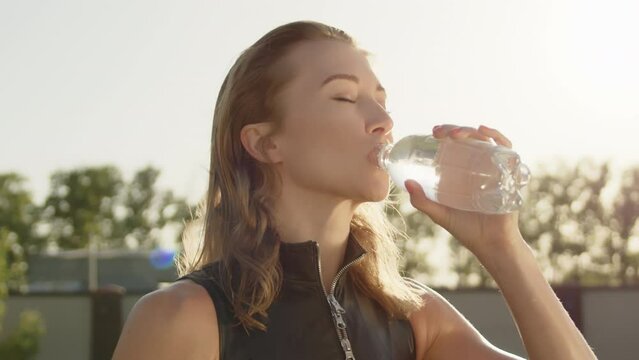 Medium shot of young cute sexy blonde drinking water from bottle after workout outdoors. Slow motion. Woman closes eyes, hair flutters in the wind. High quality 4k footage