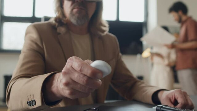 Medium close-up shot of middle-aged male product engineer in AR headset manipulating 3-dimensional virtual prototype using remote control, and colleagues discussing solar panel diagrams in background