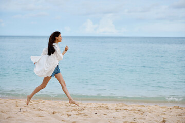Sports woman runs along the beach in summer clothes on the sand in a yellow tank top and denim shorts white shirt flying hair ocean view, beach vacation and travel