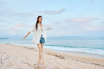 Fototapeta na wymiar Happy tanned woman in white swimsuit shirt and denim shorts walks on the beach on the sand by the ocean with wet hair after swimming, sunset light and pink clouds in Bali