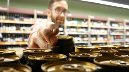 Close-up of many beer cans on a store shelf and a smiling bearded man with glasses taking one