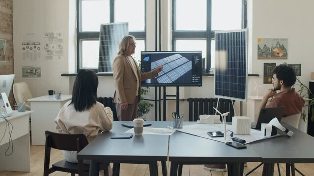 Full Shot Of Caucasian Male Engineer Presenting Solar Panel Project On Multimedia Screen To Attentive Colleagues In Office Of Renewable Energy Research Company