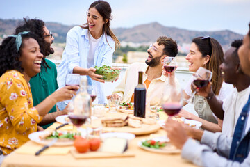 Young smiling Caucasian woman putting plate with food on table at celebration with happy friends on rooftop. Group of cheerful multiracial friends gathered for barbecue party on outdoor terrace.