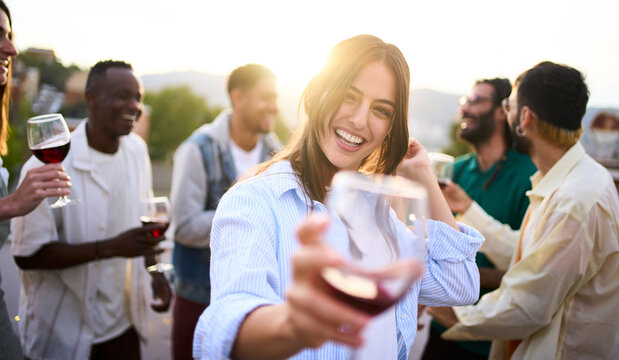 Point of view of pretty young woman toasting with glass of red wine in hand and smiling. Girl posing for photo at party with her friends outdoors at sunset. People gathered celebrating success.