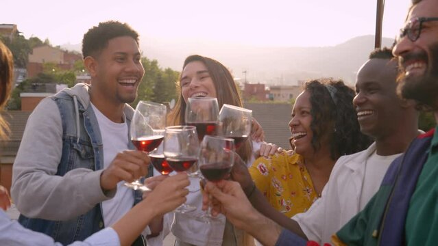 Group of multiracial friends toasting red wine and celebrating party at country house. Laughing young people hugging having fun outdoor terrace. Boys and girls enjoying free time on summer vacation.