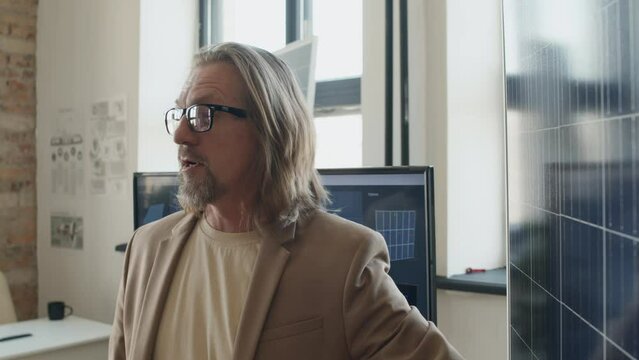 Medium close-up shot of middle-aged Caucasian male engineer in jacket and t-shirt talking about solar panel design and functions in loft style office of renewable energy development company