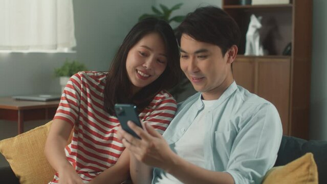 Happy young attractive Asian couple hand holing mobile phone enjoy playing social media online shopping surfing internet on smartphone smile talking feeling cheerful sit on couch at cozy living room