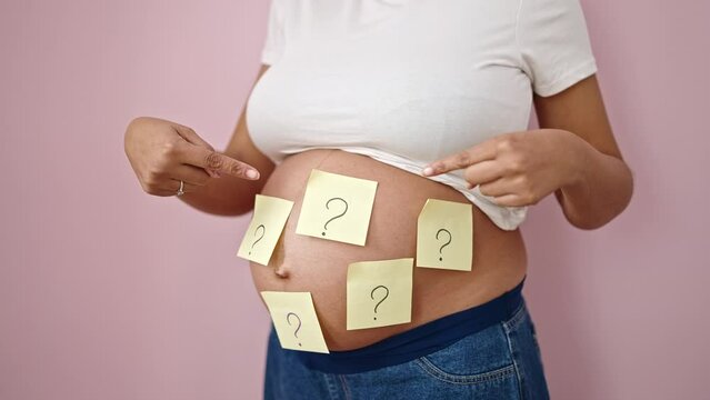 Young pregnant woman asking for beby gender pointing to question marks on belly over isolated pink background