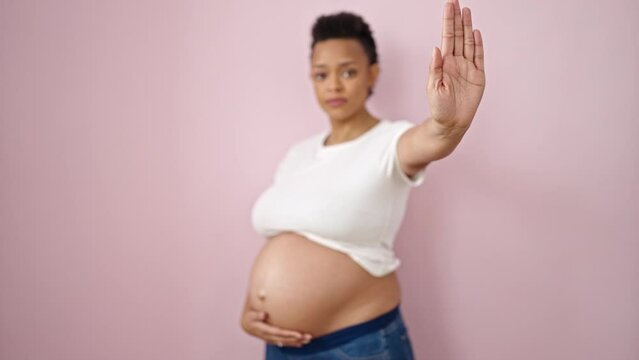 Young pregnant woman doing stop gesture with hand over isolated pink background