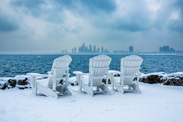 white deck chairs in winter looking out at a city sky line across open water with stormy snow filled sky