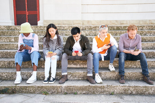 Group of serious multiracial friends using cell phone together outdoors. Young people sitting and concentrating looking at mobile. Diverse people gathered addicted to social networks.