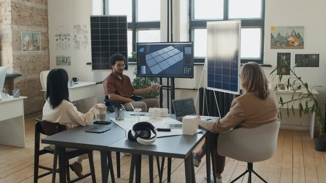 Full shot of ethnically diverse male and female engineers sitting around table at meeting in office of renewable energy company, looking at multimedia screen and discussing solar power project