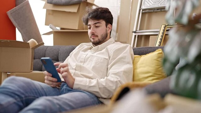 Young hispanic man using smartphone sitting on sofa at new home