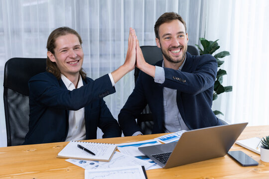 Businessmen celebrate success with joyous expressions, celebratory high-five hand gesture in office after analyzing data or achieving target. Teamwork and planning lead to successful outcome. Entity