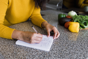 Latin adult woman cooking and writing recipe in a notebook in kitchen at home in Mexico Latin...