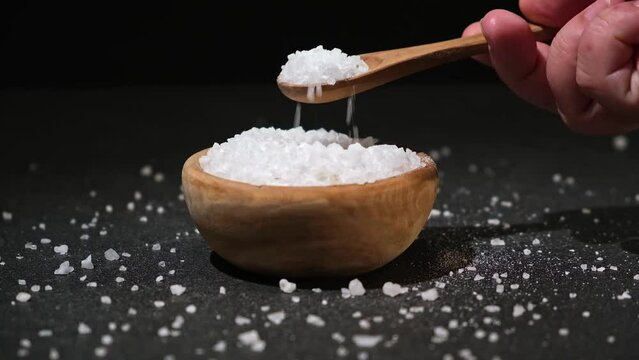 A girl dials salt into a wooden spoon from a bowl on a black background. Coarse sea salt.
