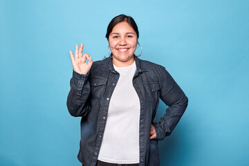Young curvy latina woman wearing denim shirt and hoop earrings, smiling showing okay sign, hand on waist, looking at camera isolated on blue background. Copy space. - obrazy, fototapety, plakaty